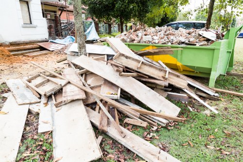 A pile of discarded wooden door frames and planks with visible textures of aged, weathered wood, light beige to greyish tones, arranged in a disorderly manner on a grassy area outside a property. Behind the debris, there is a green skip filled with bricks and rubble, situated on a paved or driveway surface. In the background, there are trees, a residential building with white walls, and some outdoor steps. The scene appears to be part of an outdoor waste clearance process, typical of rubbish removal services offered by Office Clearance Finchley, serving areas such as Finchley in north London. The overall environment indicates a garden or backyard clearance with natural daylight illuminating the setting, emphasizing the contrast between the wooden waste and the greenery of the grass and trees.
