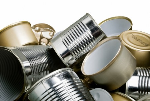 A close-up of multiple tin cans and aluminum containers piled together, with some showing their open ends revealing the inside surfaces. The cans vary in size, with some having smooth metallic finishes and others featuring ridged or embossed textures. The colours include shiny silver, gold, and metallic grey, with some cans stacked horizontally and others leaning against each other. The surface of the cans reflects light, creating highlights and shadows that accentuate their cylindrical shapes. These cans are situated on a plain background, likely in a setting associated with rubbish disposal or recycling, possibly outdoors or in a waste collection area near Finchley. The image visually represents waste material, aligning with rubbish removal services offered by Office Clearance Finchley in the local area, emphasizing the need for proper disposal or recycling of metal containers.