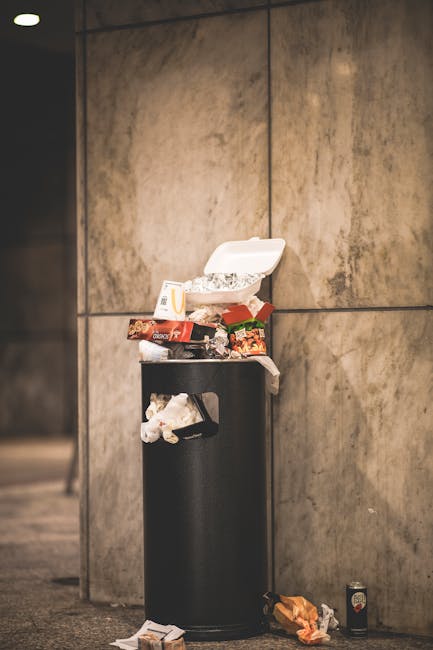 A black wheeled rubbish bin positioned on a quiet street pavement during nighttime, with a white label that reads 'ST. JOHN'S'. The bin is filled with various waste items including cardboard boxes, pl