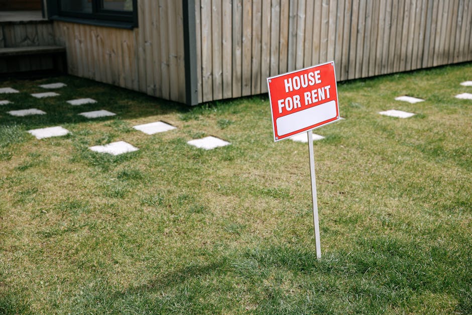 A red and white 'House For Rent' sign is mounted on a metal post in the foreground, positioned on a grassy area outside a wooden fence. Behind the sign, three individuals stand together, slightly out
